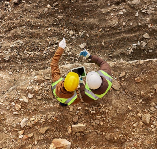 two construction workers discussing project details on a tablet in a gravel site with safety gear and helmets 3 workers construction site collaboration