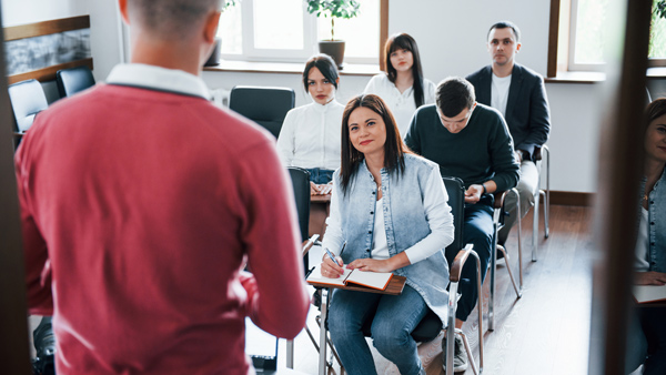 a group of people attending a presentation with one person speaking while the audience listens and takes notes in a learning environment