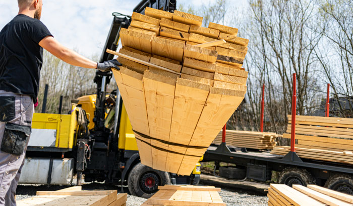 man lifting stacked wooden planks on construction site with forklift in background handling two bundles of lumber